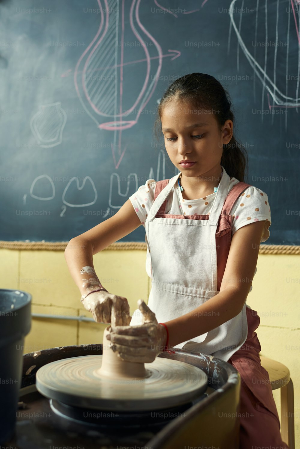 Studentessa contemporanea seduta ruotando la ruota di ceramica contro la lavagna mentre scolpendo una nuova brocca di argilla durante la lezione di artigianato