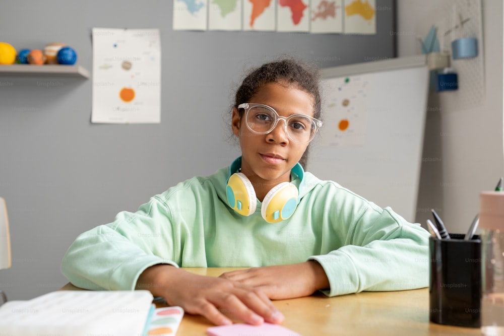 Adorable colegiala de raza mixta con anteojos y ropa informal sentada junto a su escritorio en el aula