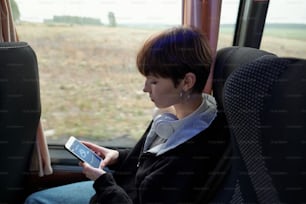 Serious Caucasian student girl with headphones around neck sitting in bus and checking weather forecast on phone