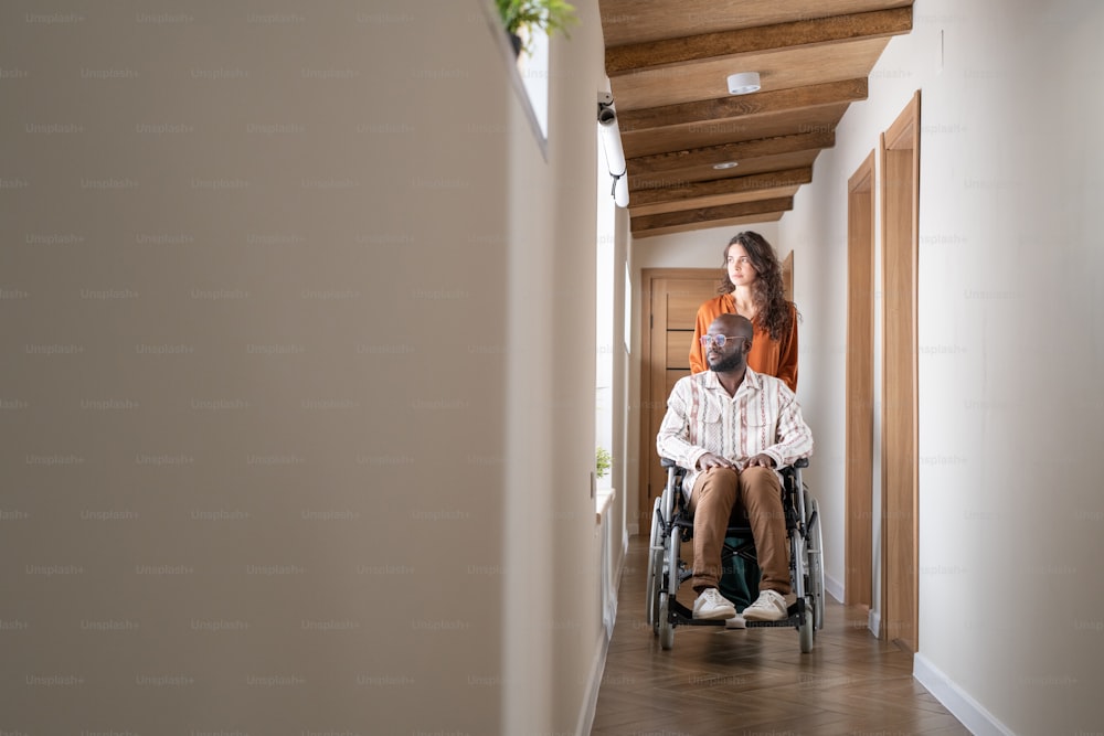 Jovem negro em trajes casuais sentado em cadeira de rodas na frente de cuidadora caucasiana ajudando-o a se mudar no apartamento
