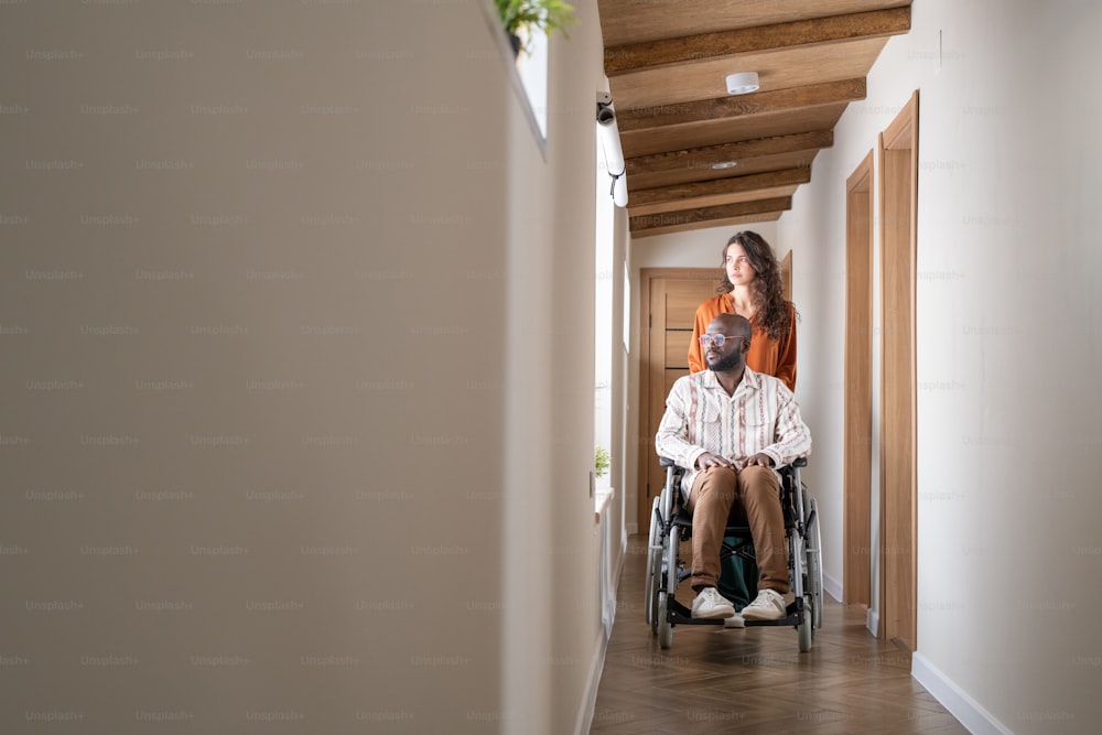 Junger schwarzer Mann in Freizeitkleidung sitzt im Rollstuhl vor kaukasischer Pflegerin und hilft ihm beim Einzug in die Wohnung
