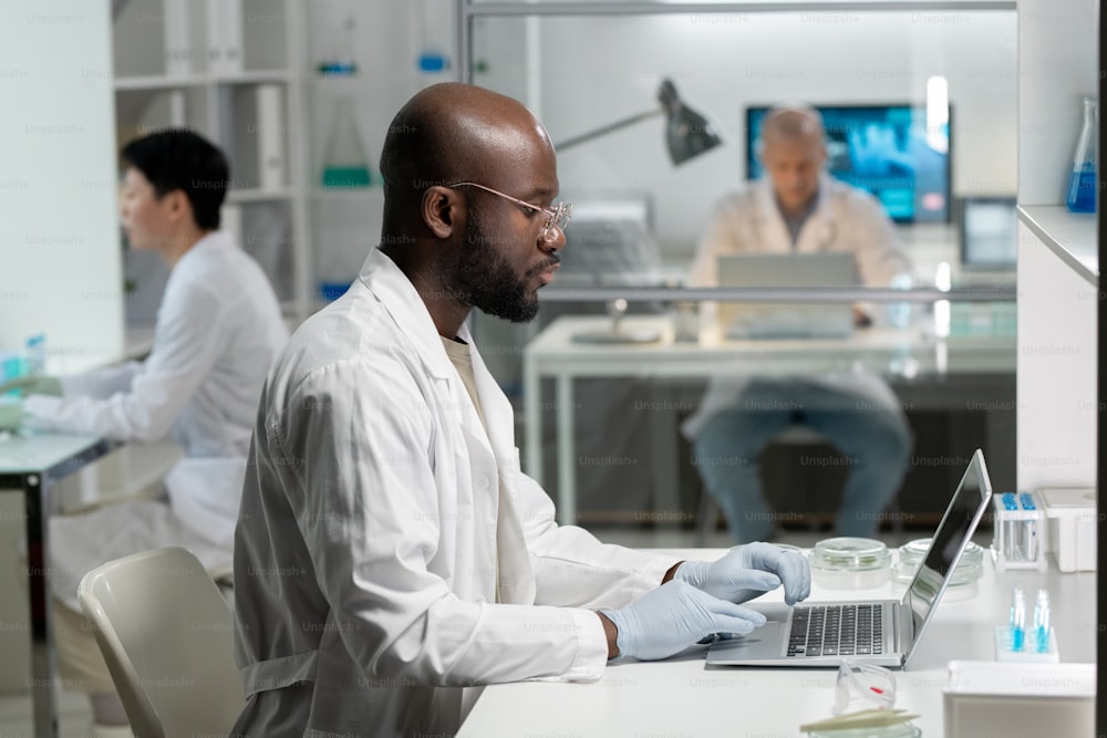 Vista lateral do jovem pesquisador afro-americano em jaleco de laboratório e luvas cirúrgicas digitando no teclado do laptop enquanto está sentado ao lado da mesa