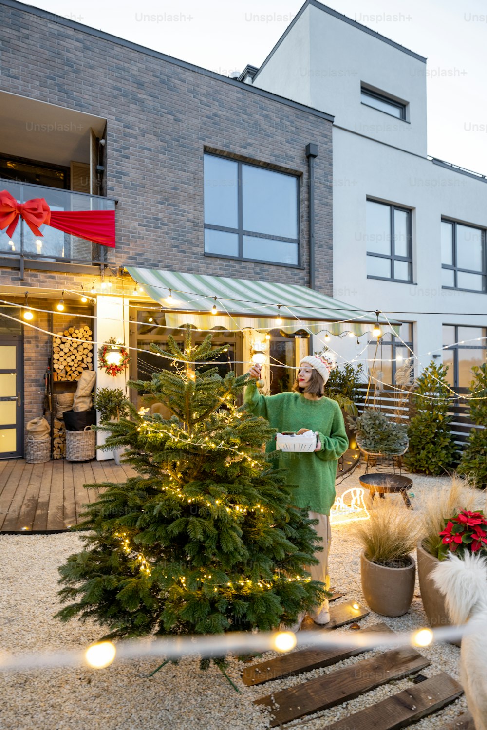 Woman decorating Christmas tree at beautiful backyard of her counry house. Concept of preparation for the winter holidays. Caucasian woman wearing green sweater and hat. Wide angle view