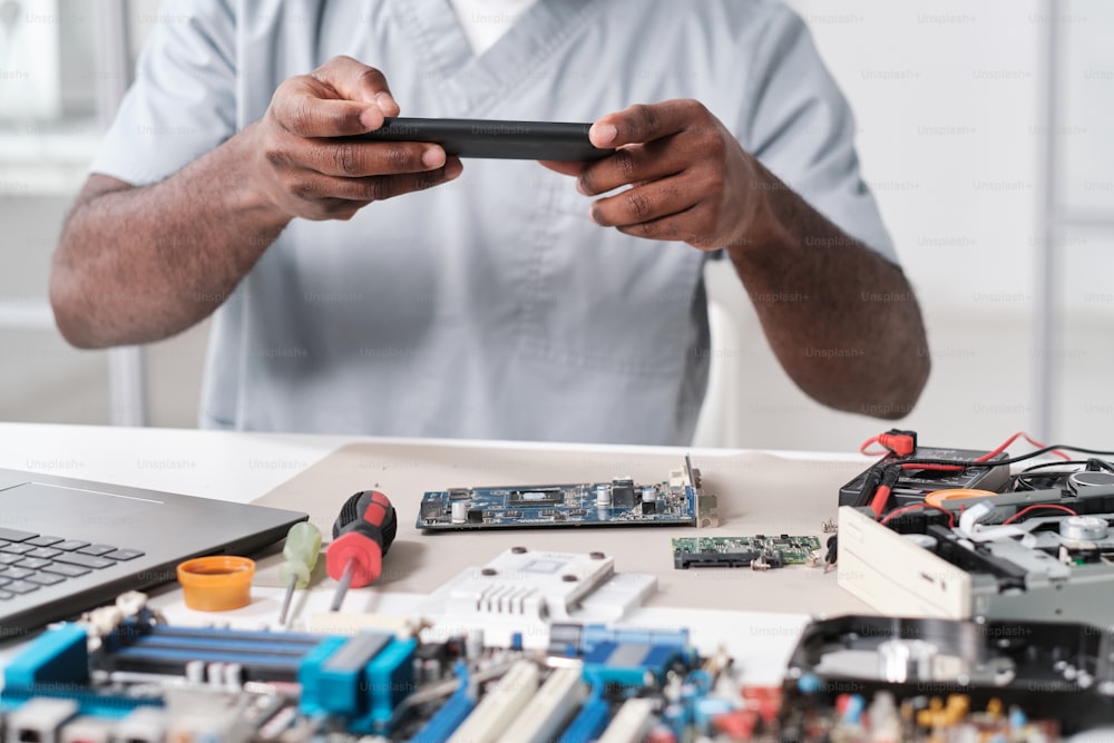 Hände eines jungen afrikanischen Reparateurs fotografieren Leiterplatte eines elektronischen Geräts, während er am Schreibtisch in der Werkstatt sitzt