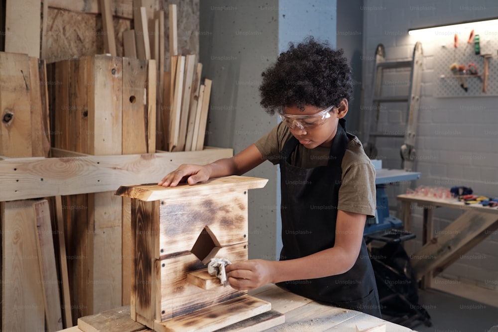 Portrait of teenage African-American boy building wooden birdhouse in carpentry workshop, copy space