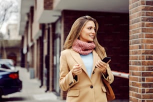 A young, happy, fashionable woman in a coat and with a scarf is walking outdoors and holding her phone in her hands. Telecommunications and global network. A woman walking outdoors.