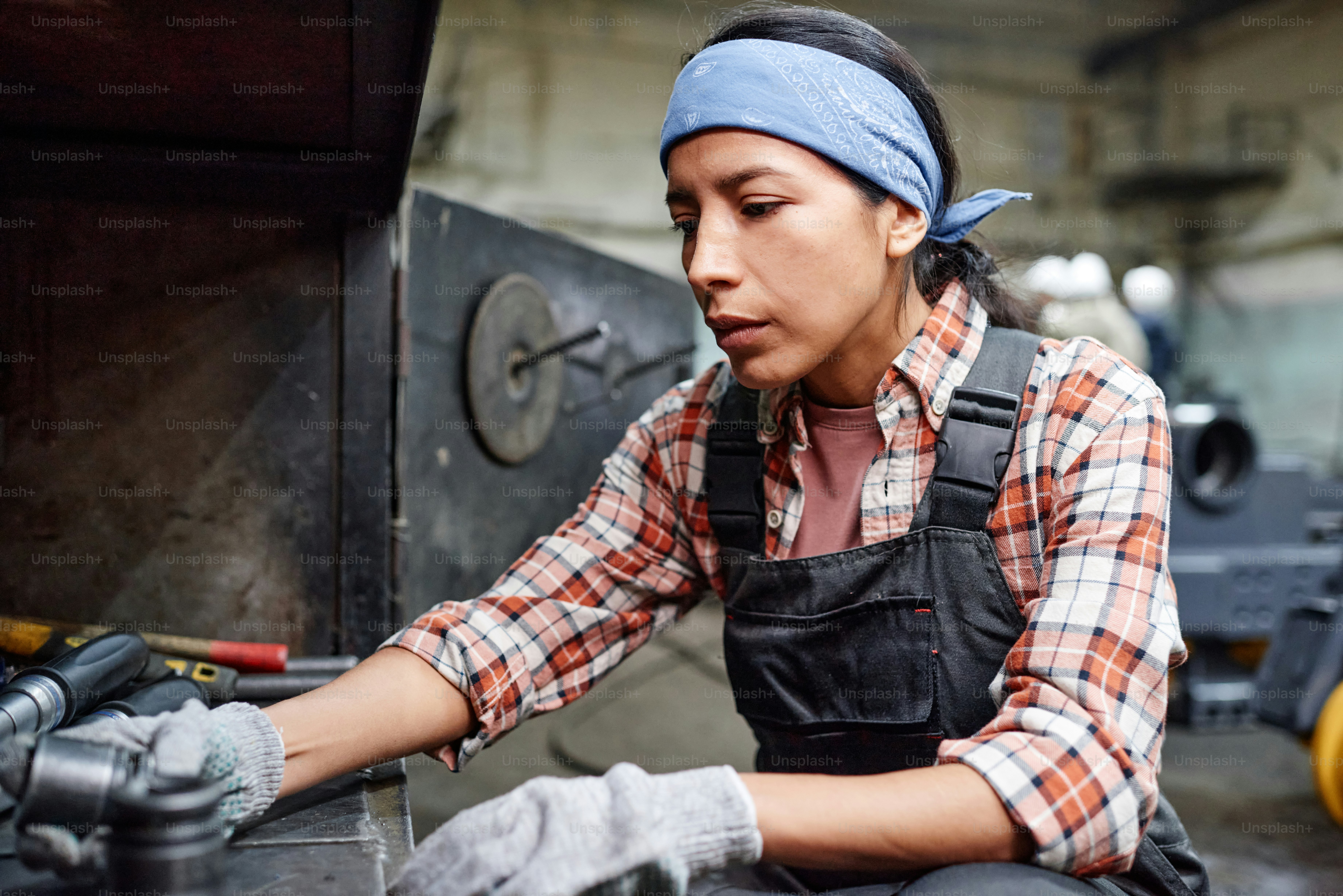 Young contemporary female in workwear choosing metallic spare parts while going to repair industrial machine in large workshop