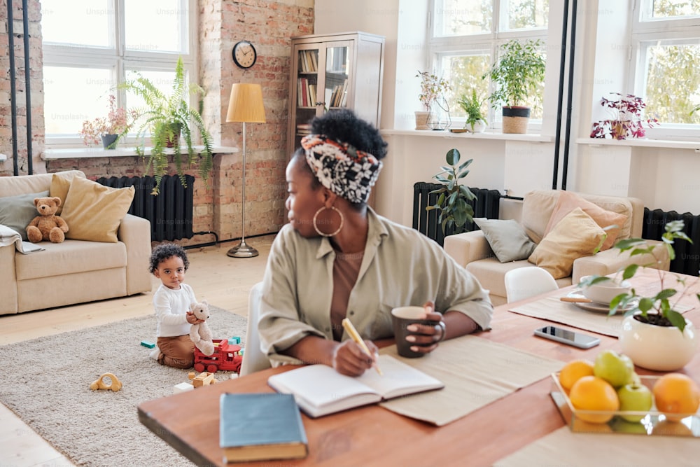 Madre negra trabajadora mirando a su hijo y tomando notas en el planificador mientras su hijo juega con un oso de juguete