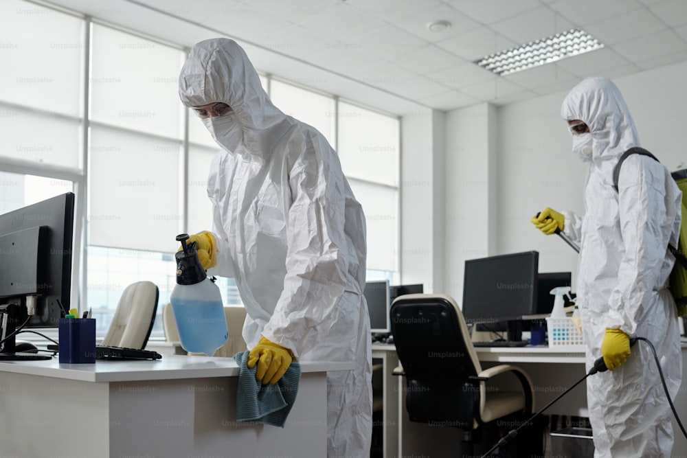 Trabalhadores do serviço de limpeza em trajes de materiais perigosos desinfetando móveis e equipamentos de informática em grandes escritórios modernos de espaço aberto