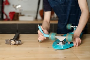 Contemporary adolescent boy with blue wrench fixing wheels of skateboard while bending over workbench with hammer