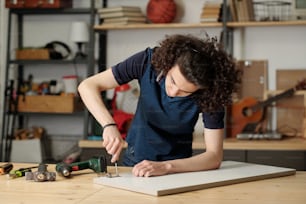 Adolescent guy in casualwear fixing door hinge with screwdreiver in garage while bending over table with plywood board