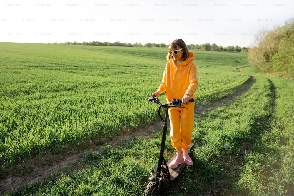 Young person in overalls riding electric scooter on green field during sunset. Concept of sustainability and eco-friendly lifestyle