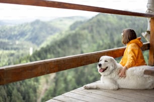 Young person in orange sport suit enjoys great mountain landscape while sitting with her dog on a wooden terrace. Concept of escape and solitude in nature and traveling with pets