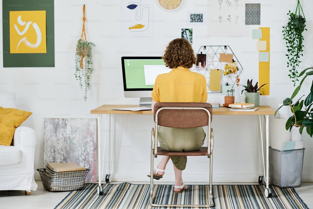 Vista traseira de jovem freelancer do sexo feminino em casualwear sentado no local de trabalho na frente do monitor do computador e criando novos sites em casa