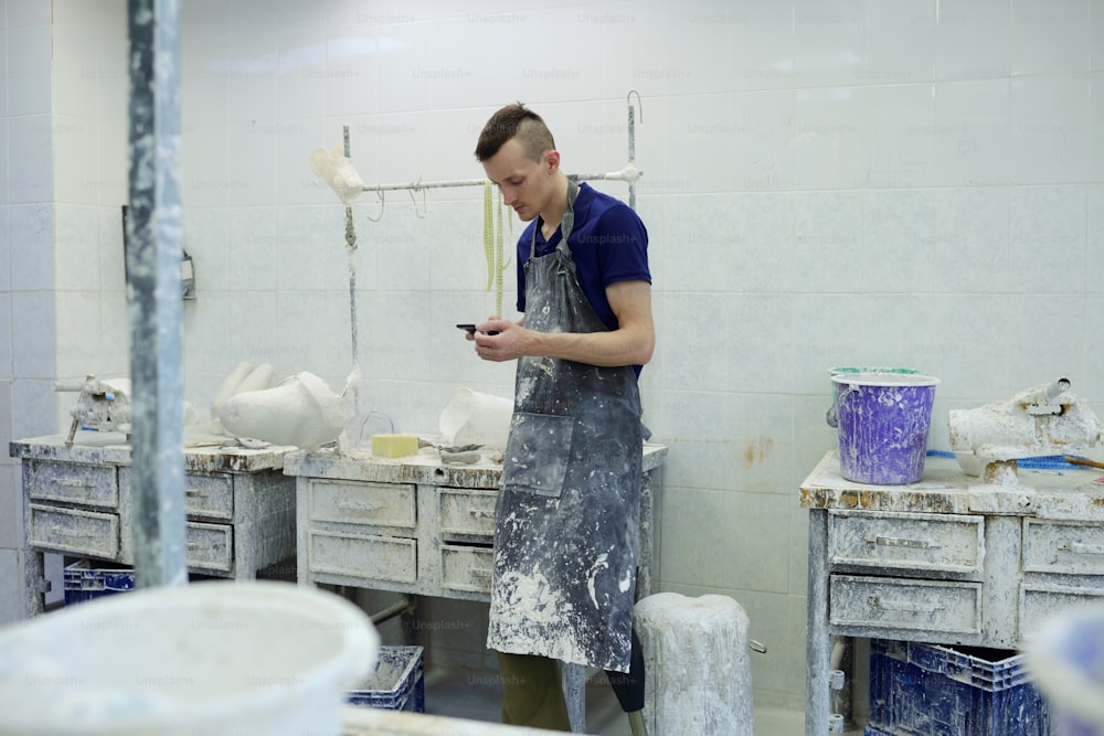 Giovane operaio di una fabbrica di protesi con disabilità che scorre nello smartphone mentre invia messaggi o sfoglia informazioni online
