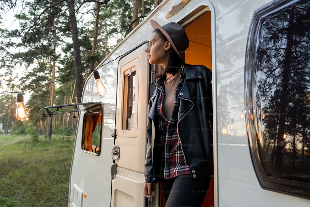 Young female traveler in hat and black leather jacket standing in open door of house on wheels in the evening