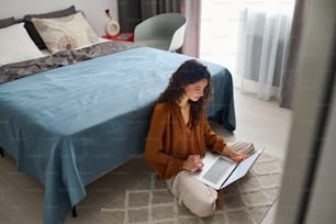 Busy young female with laptop sitting on the floor by double bed and networking or just spending free time on various websites