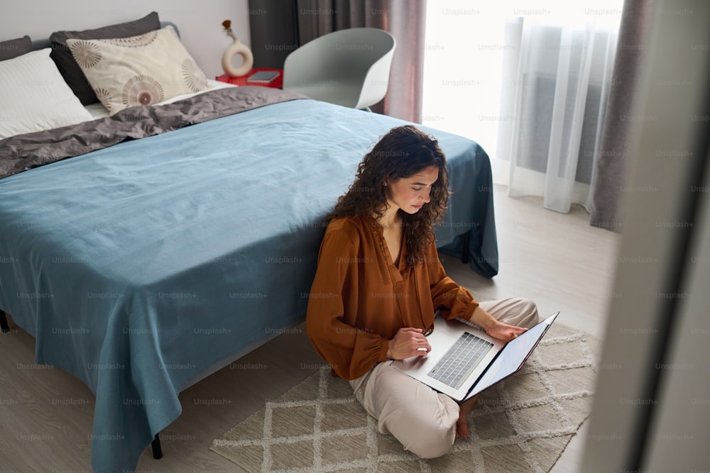 Mujer joven ocupada con computadora portátil sentada en el piso junto a la cama doble y la red o simplemente pasando el tiempo libre en varios sitios web