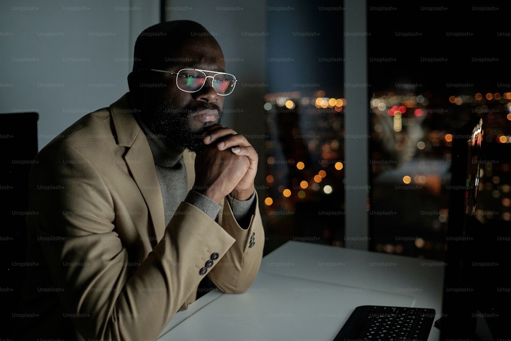 Uomo d'affari serio in abbigliamento formale che guarda lo schermo del computer mentre è seduto vicino alla scrivania in ufficio di notte
