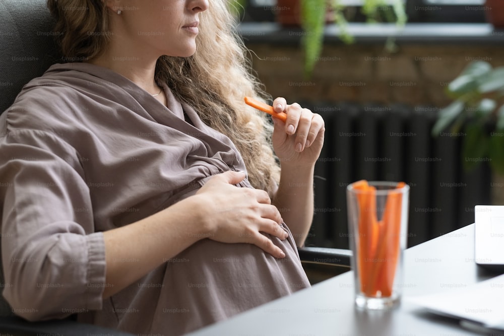 Primer plano de una mujer embarazada que se toca el vientre y come zanahorias en su lugar de trabajo en la oficina durante el almuerzo