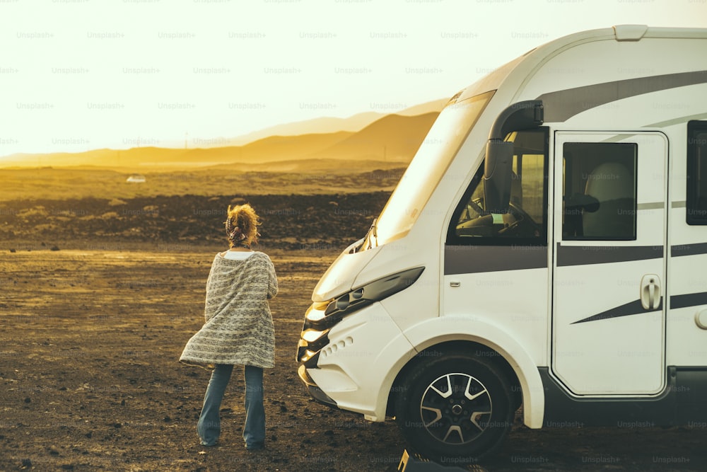 Concept of travel and freedom. Back view of standing woman admiring desert landscape near modern camper van motorhome. Alternative home lifestyle female people looking sunset free