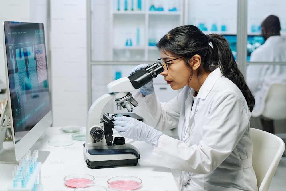 Vue latérale d’une biochimiste en blouse blanche, gants et lunettes regardant au microscope tout en étudiant le virus en laboratoire