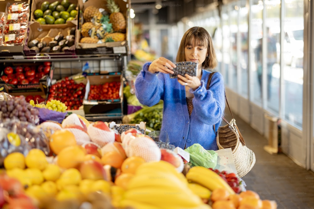 Woman choosing blueberry while shopping food at local market. Fresh fruits on the counter of market stand indoors