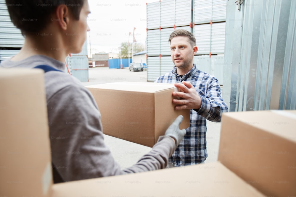 Young mover giving cardboard box to client while taking it from cargo container outdoors