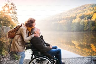 秋の美しい自然の中を散歩するアクティブなシニアカップル。早朝の湖畔で車椅子に乗った男女。