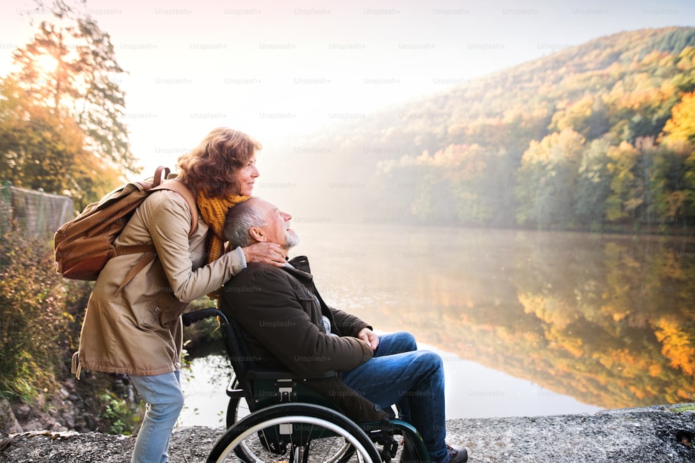 Active senior couple on a walk in a beautiful autumn nature. A woman and man in a wheelchair by the lake in the early morning.