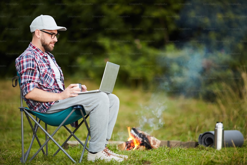 Young man sitting on chair and typing on laptop near the campfire in the forest