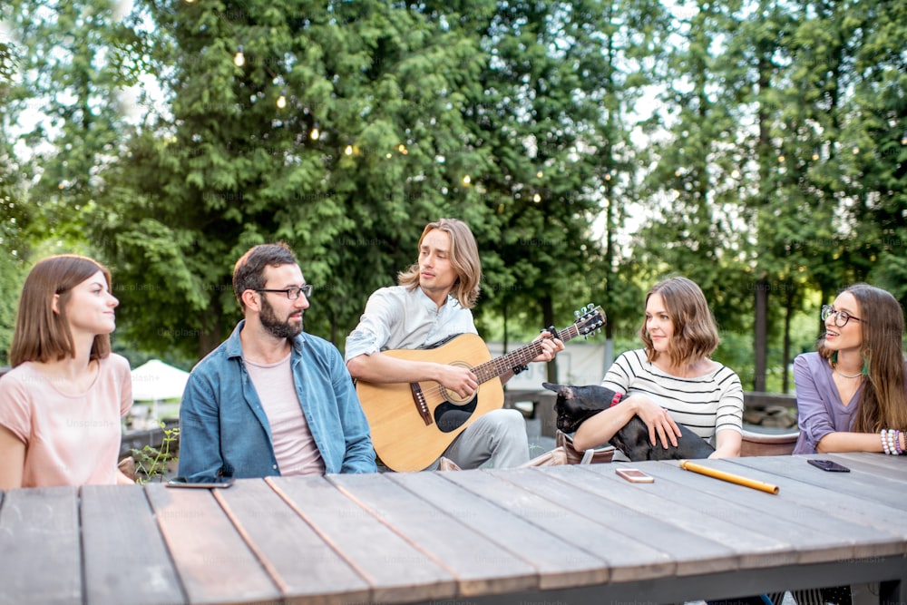 Young friends having fun together playing a guitar sitting at the table outdoors in the park