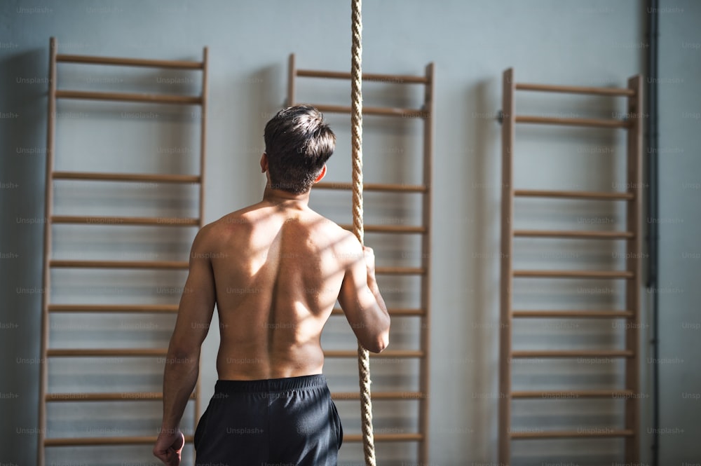 Unrecognizable fit young man in gym standing topless, holding climbing rope. Copy space. Rear view.