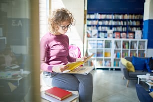 Studentessa con i capelli ricci e gli occhiali che preparano il suo rapporto, è seduta sul bordo della finestra con il libro in biblioteca