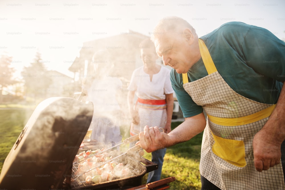 Elder happy man is checking his meat on a barbeque while his family is enjoying in a backyard