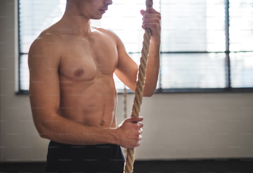 Unrecognizable fit young man in gym standing topless , holding climbing rope. Copy space.