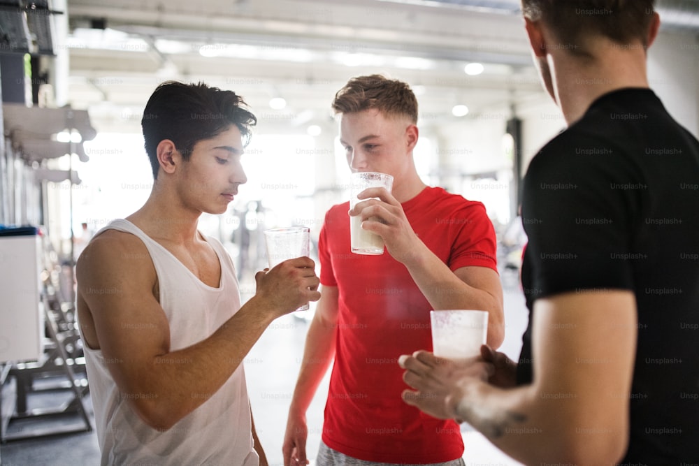 Three young fit men in gym holding glasses with protein drinks.