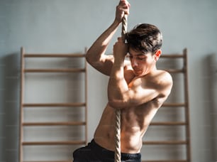 Bonito apto jovem topless homem na academia escalando uma corda. Barras de parede ao fundo.