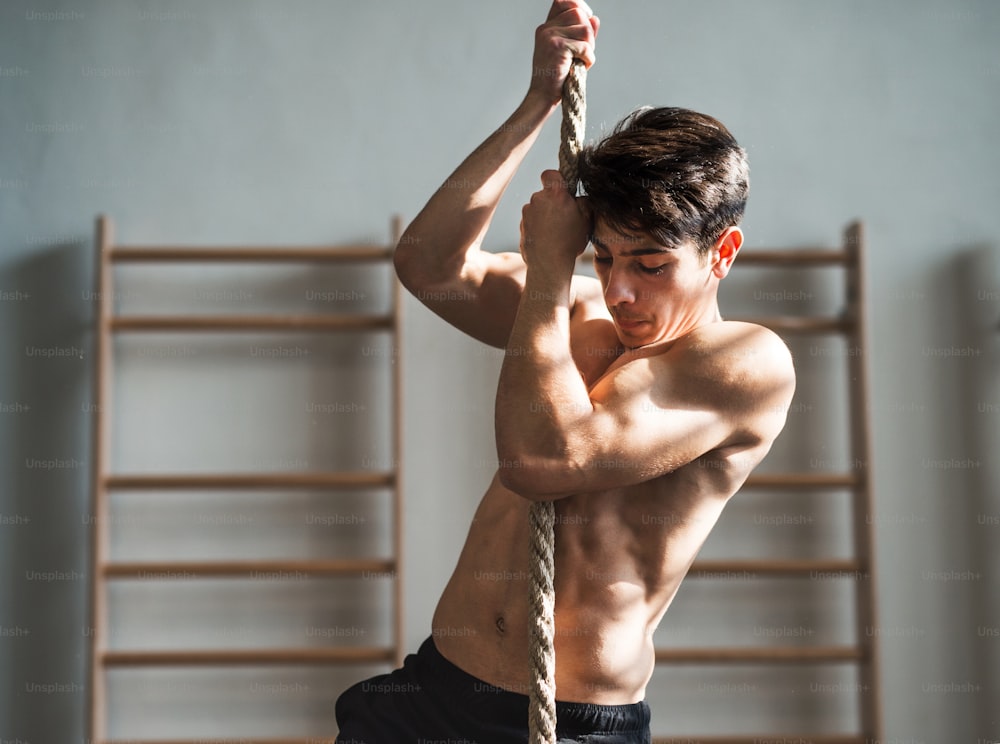 Handsome fit young topless man in gym climbing a rope. Wall-bars in the background.