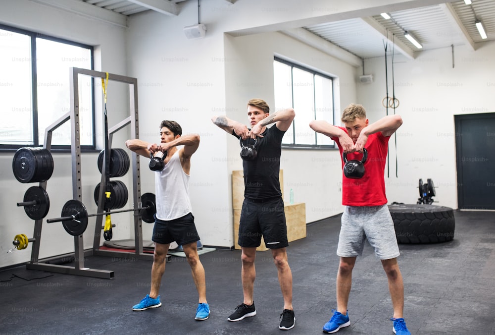 Three young fit men in gym working out, doing kettlebell swings.