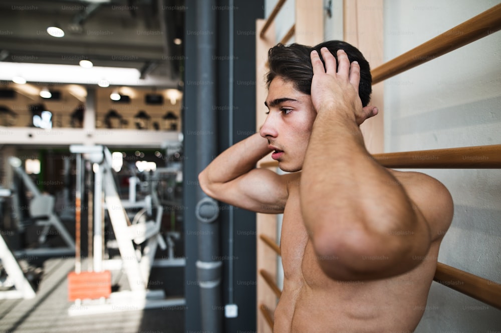 Giovane bello in forma in palestra in piedi in topless davanti alle sbarre a muro.