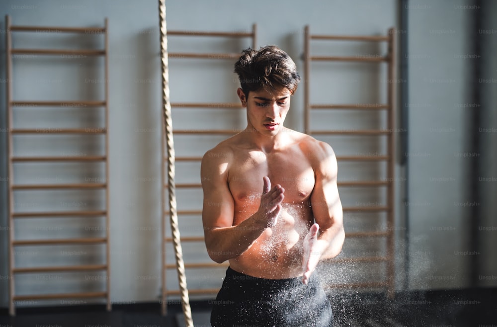 Fit young man in gym standing topless by the climbing rope , smearing his hands with magnesium.