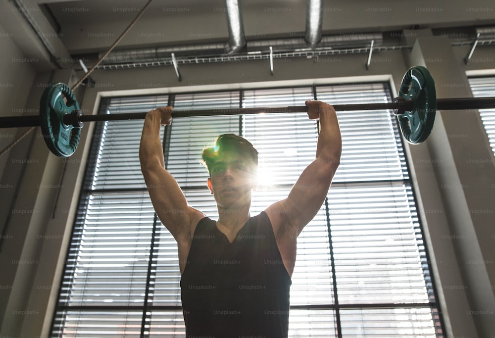 Junger, gutaussehender fitter Mann im Fitnessstudio trainieren, Hantelstange heben, Muskeln spielen lassen.