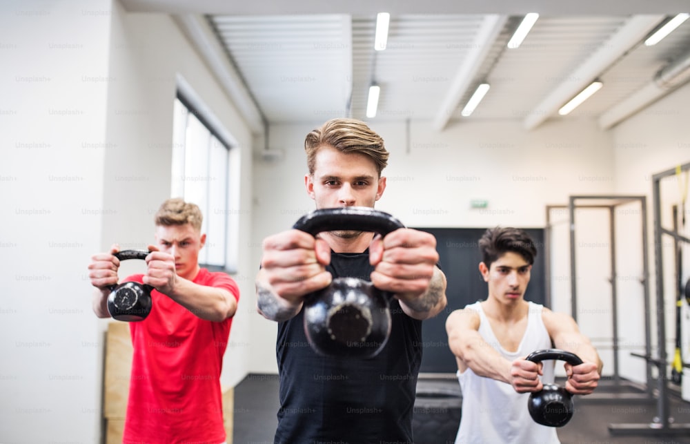 Three young fit men in gym working out, doing kettlebell swings.