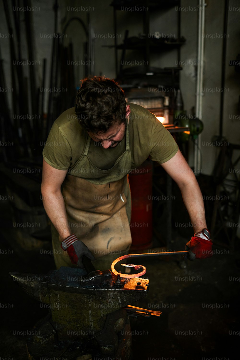 Young blacksmith in workwear leaning over anvil and working with hammer and molten metal workpiece