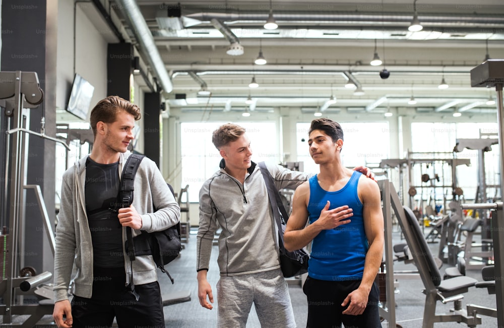 Junge männliche Freunde mit Taschen im modernen Fitnessstudio, die sich begrüßen.
