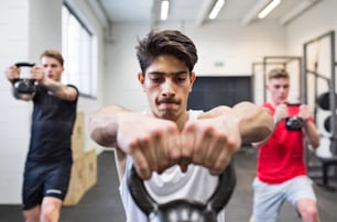 Three young fit men in gym gym working out, doing kettlebell swings.