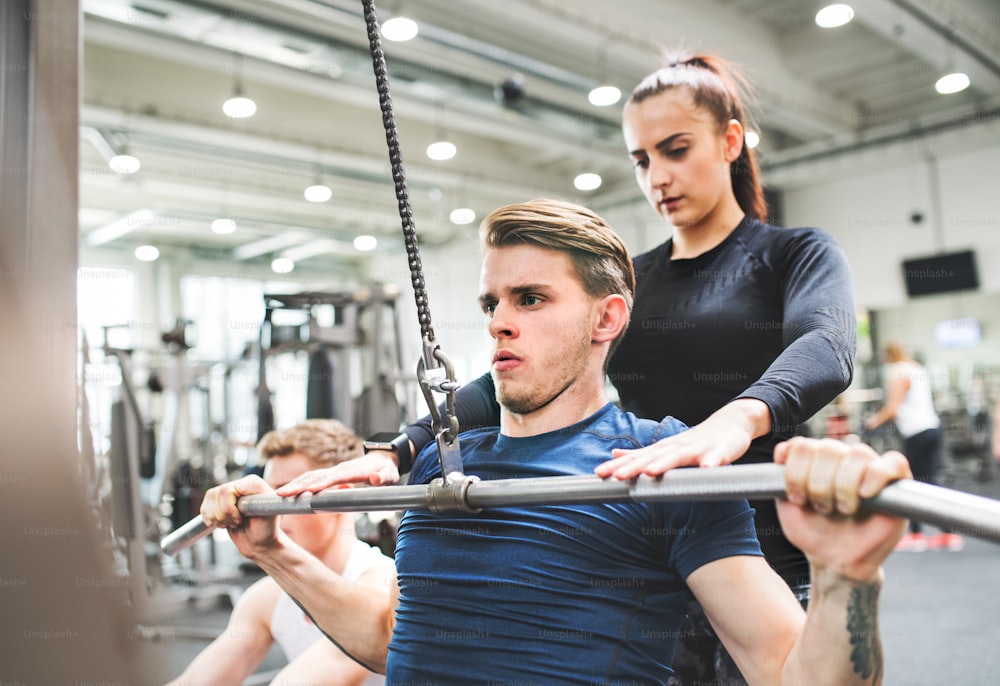 Young handsome fit man working out on pull-down machine in gym a personal trainer standing behind, helping him. Bodybuilder exercising with cable weight machine.