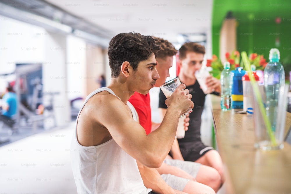 Tre giovani uomini in forma in palestra seduti e tenendo bicchieri con bevande proteiche.