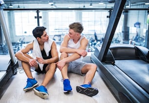 Two young fit men in gym gym sitting and resting after an exercise, talking.